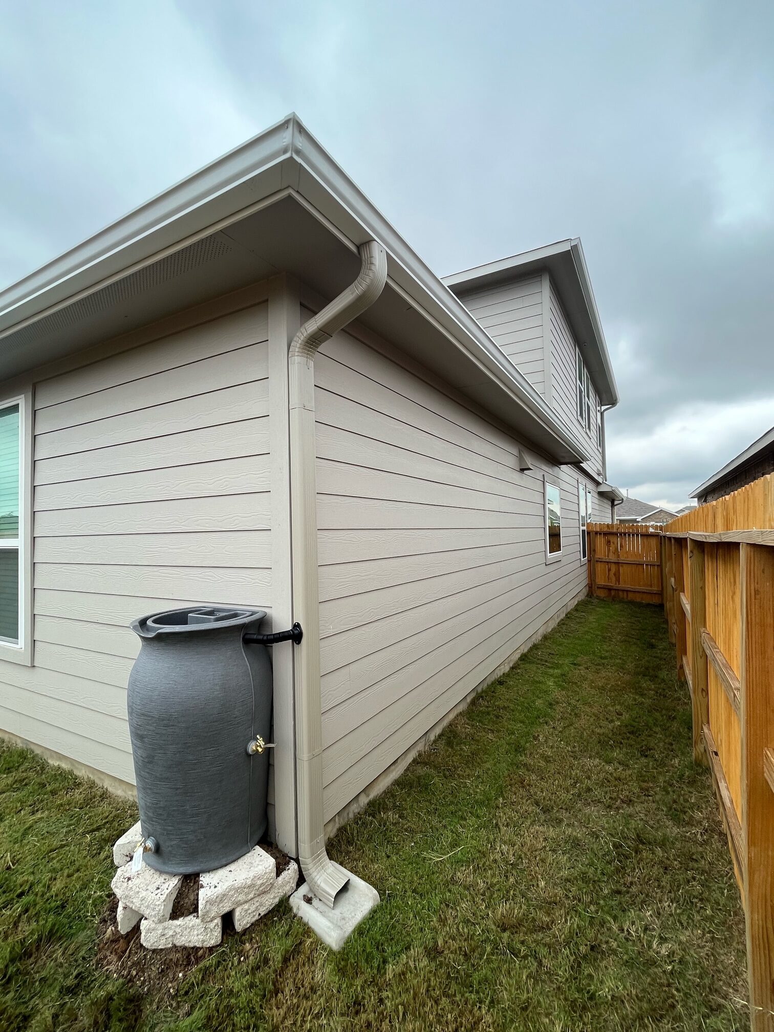 Gray rain barrel fed by a seamless downspout and set on a paver stand beside a house